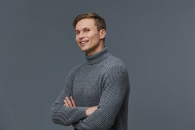Açık kahverengi saçlı, gri balıkçı yaka kazaklı, gri arka planda poz veren mutlu bir adam. Günlük kıyafet, pozitif ruh hali, kapalı stüdyo çekimi rahatlama ve...