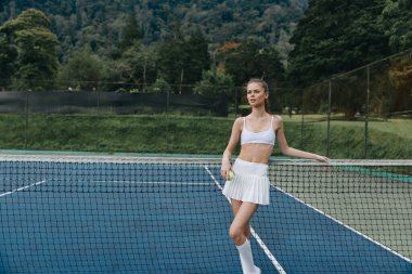 Beyazlar içinde kendine güvenen genç bayan tenisçi tenis kortunda duruyor, somutlaşmış atletizm ve zarafet dolu çevre, spor markalaşması için mükemmel.