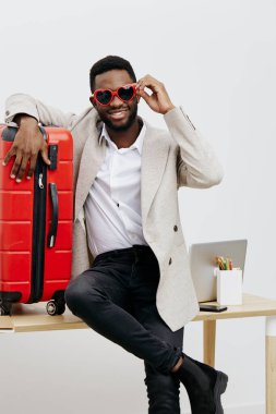 Kalp şeklinde güneş gözlüğü takan, bavula yaslanan, dizüstü bilgisayarı ve kırtasiyesi olan, seyahat veya seyahat için heyecanını ifade eden, modern gündelik görünüş, neşeli ruh hali, seyahat