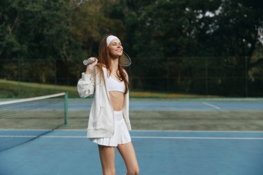 Güneşli bir günde tenisin keyfini çıkaran beyaz elbiseli mutlu genç bayan sporcu, canlı bir kort geçmişinde sağlıklı yaşam tarzı ve sportmenliğin vücut bulmuş hali.