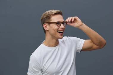 Kısa kahverengi saçlı ve gözlüklü mutlu genç adam, gözlüğünü tutarken geniş bir gülümseme, günlük beyaz tişört, düz gri arka plan, rahat ruh hali, stüdyo portresi, yaklaşılabilir ve