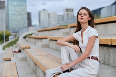 Şık bir iş kıyafeti giymiş dengeli bir kadın, açık tribünlere oturmuş, şehrin modern bir arka planına karşı, özgüven ve profesyonelliği somutlaştırıyor..