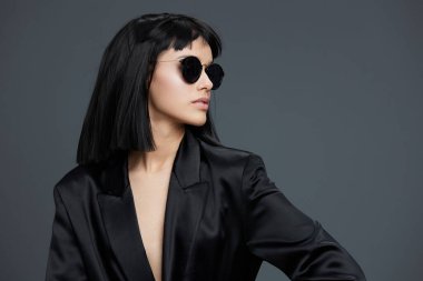 Güneş gözlüğü takan güzel Asyalı bir kadın ve siyah ceketli, gri bir arka plana karşı güvenle poz veren, zarafet saçan ve modern tarzda