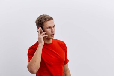 Beyaz arka planda izole edilmiş kırmızı tişörtlü, kırmızı tişörtlü, cep telefonuyla konuşan beyaz bir genç. Mobil teknolojiyle modern iletişim ve dijital göçebe yaşam tarzı kavramı