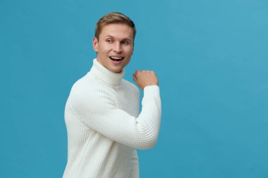 Beyaz süveter giymiş gülümseyen bir adam mavi arka planda omzunun üstünden bakıyor. Genç, beyaz bir adam günlük kıyafetlere duyduğu güveni ifade ediyor. Konsept: Uzak çalışma ile dijital göçebe yaşam tarzı