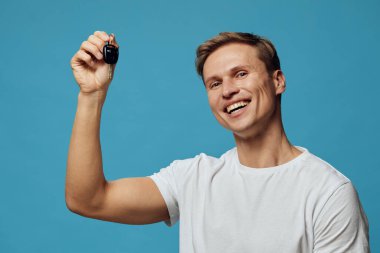 Kısa kahverengi saçlı ve günlük beyaz tişörtlü mutlu genç adam mavi arka planda araba anahtarını sunuyor. Heyecanlı duygular, kutlamalar, özgüven, başarı, yaşam tarzı