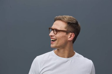 Kısa kahverengi saçlı, gözlüklü, beyaz bir tişört giyen, gülümseyen ve sade gri bir stüdyo geçmişi olan mutlu bir adam. Kavram: neşeli, gündelik, pozitif ruh hali.