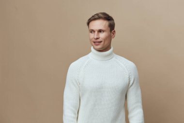 Beyaz balıkçı yaka kazak giyen, gülümseyen ve stüdyo çekiminde bej bir arka plana bakan dost canlısı bir adam. Sıcak, rahat kıyafetler, günlük tarz, neşeli ruh hali.