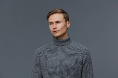 Gri balıkçı yaka kazak giyen düşünceli genç adam arka plandaki gri stüdyo resmine yan bakıyor. Sıradan moda kıyafetleri, akıl sağlığı ve diğer aletler modern yaşam tarzını ön plana çıkarıyor.