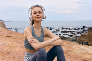 Kulaklık takan mutlu genç bir kadın, şık gri bir spor kıyafeti giyiyor, plajda dinleniyor sakin okyanus manzarası rahatlatıcı bir atmosfer yaratıyor.