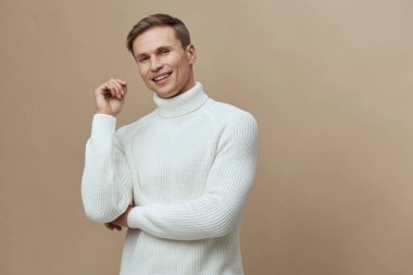 Beyaz balıkçı yaka kazak giyen gülümseyen bir adam kameraya bakıyor. Neşeli yüz ifadesi olan kolları çaprazlanmış. Sade bej arka plan stüdyo resminde dijital detoks ve akıl sağlığını temsil eden izole edilmiş.