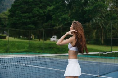 Beyaz tenis kıyafeti giymiş atletik bir kadın, yemyeşil ile çevrili bir tenis kortunda, canlı ve sağlıklı bir yaşam tarzı yayarak spor temalı içerik için ideal.