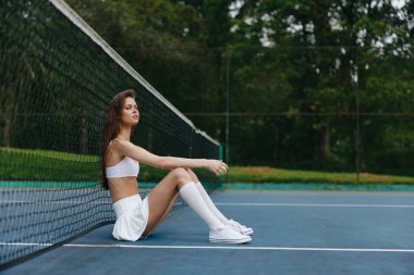 Tenis kortunda oturan beyaz spor kıyafetli atletik genç kadın, canlı yeşil bir zemin içinde kendine güven ve sükuneti yansıtıyor, sağlıklı bir yaşam tarzı görüntüsü için mükemmel.