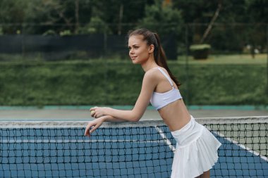 Beyaz spor giyimli kendine güvenen genç bir kadın tenis ağına yaslanıyor, güneşli açık hava oyunlarının keyfini çıkarıyor spor yaşam tarzı, fitness ve yaz temalı projeler için ideal.