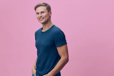 Mavi tişörtlü mutlu genç adam pembe arka planda duruyor, kendine güvenen, rahat poz veren, parlak stüdyo ortamında. Yaşam tarzı, pozitiflik ve sağlık konularına uygundur..