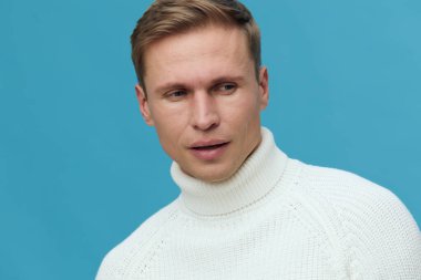 Beyaz balıkçı yaka kazak giyen beyaz tenli genç adam düşünceli görünüyor ve mavi arka planda ciddi bir yüz ifadesi var. Sakin bir stüdyo portresi ve temiz bir zihinsel sağlık ve cihazlar konsepti.