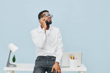Kıvırcık saçlı ve gözlüklü genç bir adam cep telefonuyla konuşuyor, beyaz gömlek ve siyah kot pantolon giyiyor, dizüstü bilgisayarı ve ofis aksesuarları var, mavi duvar