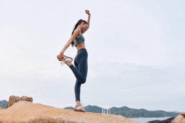 Atletik bir Asyalı kadın, gün doğumunda kayalık bir yüzeyde, sağlıklı bir yaşam tarzı ve şık bir spor kıyafetinin hedeflerini somutlaştırıyor.