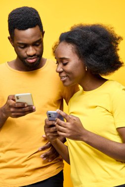 Akıllı telefonlarıyla meşgul genç bir siyah çift, modern iletişim ve samimiyeti gösteren, parlak sarı bir arka plana karşı mutluluk ve bağlantı sergiliyorlar.