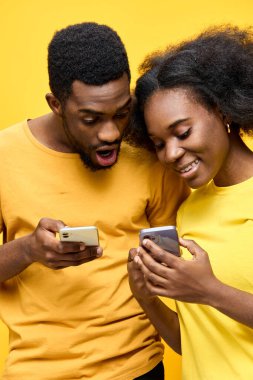 Genç bir Afrikalı çift, modern ilişkileri ve teknolojiyi temsil eden canlı sarı bir zemin üzerine kurulmuş akıllı telefonlarına bakarken meşgul ve gülümsüyor.