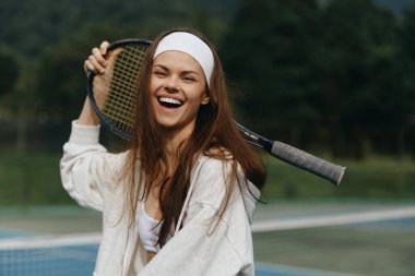 Spor kıyafetli neşeli genç bir kadın tenis raketiyle, canlı bir tenis kortunda mutluluk ve enerji saçıyor spor ve spor temaları için mükemmel.