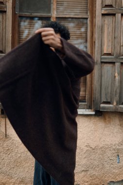 Moda adamı, omuzlarında battaniyeyle binanın önünde duruyor. Sanki bir şey saklıyormuş gibi ellerini havaya kaldırıyor..