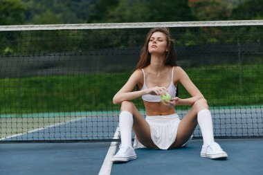 Spor giyimli genç bir bayan tenis kortunda oturmuş, sağlıklı bir atıştırmalığın tadını çıkarıyor. Canlı yeşil bir arka plana karşı güven ve rahatlama sergiliyor.