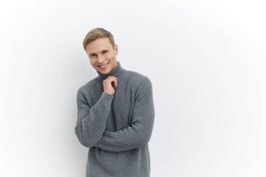 Gri örgü süveter giyen gülümseyen, beyaz arka planda tek başına duran beyaz tenli genç bir adam. Elle düşünen ve uzaktan dijital göçebe yaşam tarzı kavramı üzerinde çalışan çenesini tut.