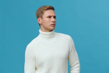 Beyaz balıkçı yaka kazaklı bir erkek model mavi bir zemine karşı düşünceli bir şekilde poz veriyor. Minimalist stüdyo ayarı tarafsız bir ifade ve doğal bir ifadeyle günlük kışlık kıyafetleri ön plana çıkarıyor.