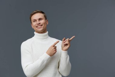 Kısa kahverengi saçlı, rahat beyaz balıkçı yaka kazak giyen neşeli genç adam, sagını göstererek mutluluğu ifade ediyor, geniş bir gülümsemeyle, düz gri bir stüdyoya yaslanıyor.
