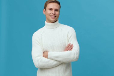 Beyaz balıkçı yaka kazak giyen gülümseyen genç bir adam kollarını kavuşturmuş mavi arka plandaki kameraya bakarak kendine güvenini ve sakinliğini gösteriyor. Görüntü, zihinsel sağlık farkındalığını ve