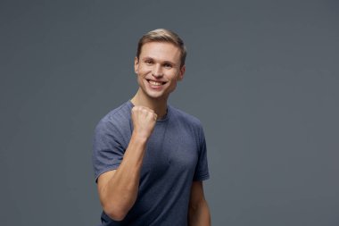 Açık sarı saçlı, sıradan mavi tişörtlü, gri stüdyo arka planında poz veren neşeli gülümseyen adam. Yakın plan portre mutluluğu, iyimserliği ve kendine güveni yakalıyor, pozitife uygun.
