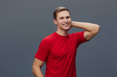 Açık tenli, kısa saçlı, sıradan kırmızı tişörtlü, kendine güvenen ve bir eli başının arkasında, düz mavi bir arka planda duran mutlu bir genç adam. Kavram: pozitiflik