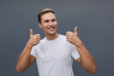Kısa kahverengi saçlı, pozitifliğini ifade eden mutlu bir adam. Başparmak kaldırma hareketi yapıyor, günlük beyaz tişört giyiyor, geniş bir gülümseme sergiliyor, yaşam tarzı ve mutluluk temaları için gri arka planda izole edilmiş.