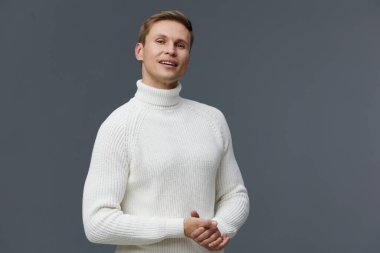 Açık kahverengi saçlı, açık tenli, balıkçı yaka kazak giyen neşeli genç bir adam, basit bir gri zemine karşı güvenle poz veriyor. Konsept: gündelik tarz ve pozitif ruh hali.