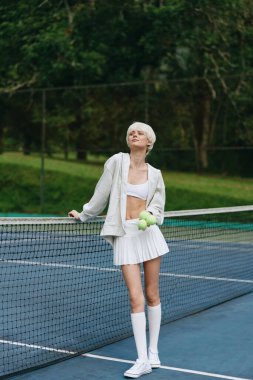 Kendine güveni tam genç bir kadın, açık renkli giyiniyor, yeşil bir geçmişi olan bir tenis kortunda neşeli poz veriyor, yaz tatili ve atletizmi yansıtıyor, tenis, spor, yaz, oyunculuk.