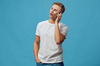 Sarı saçlı, sıradan beyaz tişört ve kot giyen, telefonu kulağına dayayan ve konuşan arkadaş canlısı genç bir adam. Mavi stüdyo, iletişimi vurgulayan temiz ve modern bir görünüm yaratıyor.