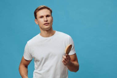 Beyaz t-shirt giyen beyaz bir genç adam mavi arka planda duran ahşap bir saç fırçası tutuyor. Kameraya bak. Ciddi ruh hali, zihinsel sağlık araçları, konsept destek ve kişisel bakım.