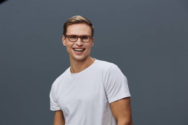 Kısa kahverengi saçlı, gözlüklü, beyaz tişörtlü ve gri arka planda poz veren mutlu bir adam. Görüntü, dost canlısı ve cana yakın bir ruh hali yakalıyor. Yaşam tarzı için mükemmel.