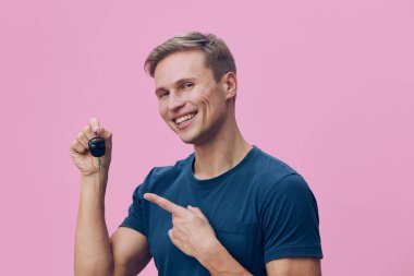 Kısa sarı saçlı, gündelik koyu mavi tişörtlü, küçük siyah bir nesneyi tutarken geniş bir gülümseme gösteren mutlu genç bir adam, pembe bir arka plana karşı durarak, ilgi uyandıran