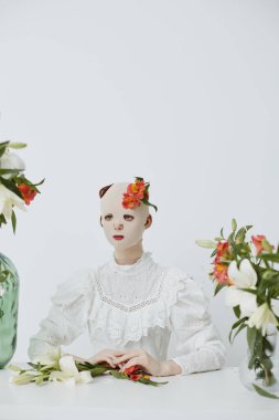 Bu sanatsal natürmort, canlı bir çiçek başlığıyla süslenmiş, eski beyaz dantel bir elbise giymiş, dingin beyaz bir alanda, taze çiçeklerle süslenmiş bir mankeni sergiliyor.
