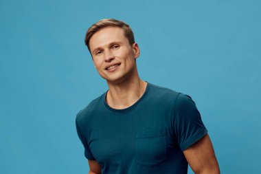 Açık tenli, kısa kahverengi saçlı, günlük mavi tişörtlü, rahat bir pozla içten bir şekilde gülümseyen, turkuaz bir stüdyonun önünde duran neşeli bir genç. Konsept: pozitif ruh hali