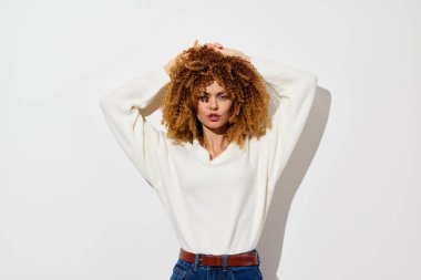 Havalı kıvırcık saçlı, rahat beyaz bir kazak ve yüksek bel kotuyla şık genç bir manken. Minimalist beyaz bir arka plana karşı cesur bir poz veriyor. Kendine güveni ve cazibesi var.