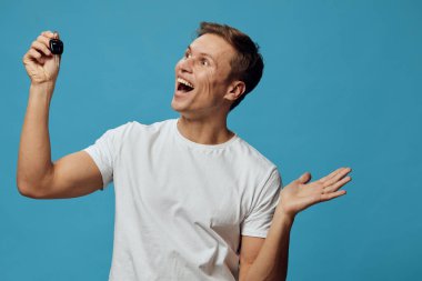 Kısa saçlı, beyaz tişörtlü mutlu bir adam, kamerayı bir eliyle tutuyor ve diğer eliyle işaret ediyor, geniş bir şekilde gülümsüyor, canlı mavi arka plan, günlük tarz, neşeli atmosfer, stüdyo