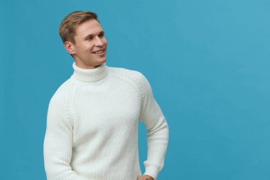 Gülümseyen beyaz balıkçı yaka kazak giyen beyaz tenli genç adam mavi arka planda rahat bir yüz ifadesiyle yan tarafa bakıyor. Kışlık rahat bir iç mekan portresi dijital göçebe üzerine odaklandı