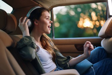 Mutlu genç bir kadın arabada şarkı söylerken, kulaklık takıyor ve günbatımında güzel bir arka plan, neşe ve özgürlüğü yansıtıyor..