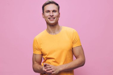 Kısa saçlı, sarı tişörtlü ve özgüvenle gülümseyen mutlu genç bir adam stüdyo ortamında pembe bir arka plana karşı duruyor. Rahat ve dostane bir ruh hali, sıradan moda, yaşam tarzı imajı.