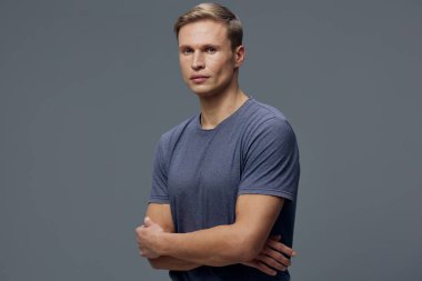 ... kolları çapraz duran ve gri arka planda kameraya bakan sıradan mavi tişörtlü, ciddi bir beyaz adam. Portre sakin ve odaklanmayı yansıtıyor, dijital detoks ve