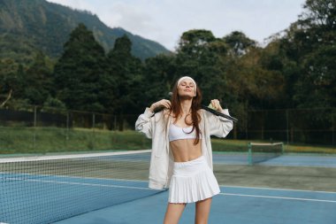 Canlı genç bayan tenisçi, beyaz etekli ve ekinlerle parıldayan neşe ve enerji saçan bir tenisçiyle dolu bir tenis kortunda, yemyeşil bir alanda raket tutarken.