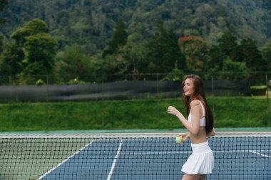 Beyaz kıyafetli enerjik bayan sporcu dışarıda tenis oynuyor, arkasında güzel yeşil bir manzara ile neşe ve sağlık yayıyor, spor ve sağlık kavramları için ideal.